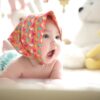 Baby lying on bed wearing a colorful bonnet, surrounded by soft toys in Pakistan including yellow and white teddy bears, evoking comfort and emotional connection.
