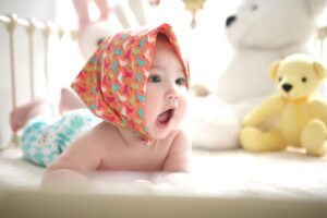Baby lying on bed wearing a colorful bonnet, surrounded by soft toys in Pakistan including yellow and white teddy bears, evoking comfort and emotional connection.